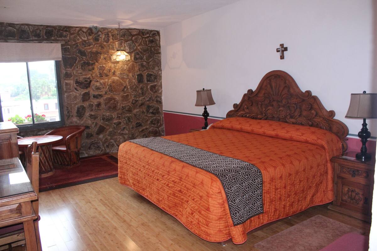 A bedroom with a stone wall, a wooden bed, a desk with chairs, and a window with a view.
