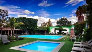 A swimming pool with two sections, surrounded by green artificial grass, lounge chairs, and a building with a green facade.