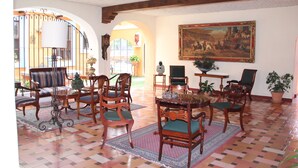 A spacious room with a large dining table surrounded by wooden chairs, a large painting on the wall, and a view of the outside through an arched doorway.