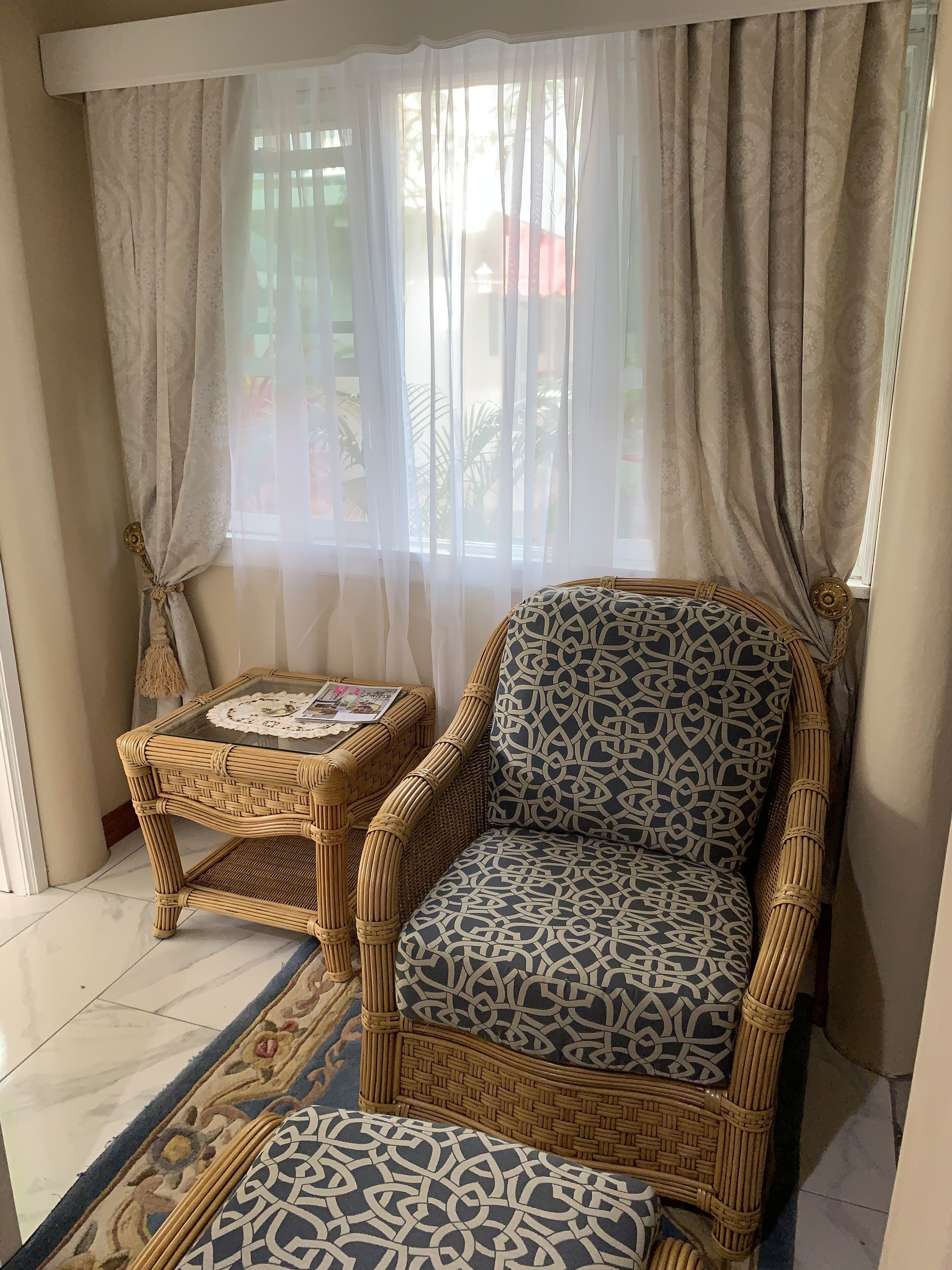 Un sillón de mimbre con un cojín estampado, una mesita de mimbre con un bordado de encaje, y una alfombra estampada sobre un suelo de baldosas.