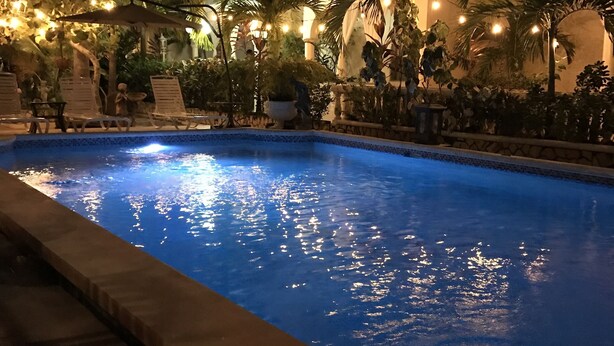 Zona de piscina nocturna con una piscina azul cristalina, rodeada de palmeras e iluminación ambiental cálida.