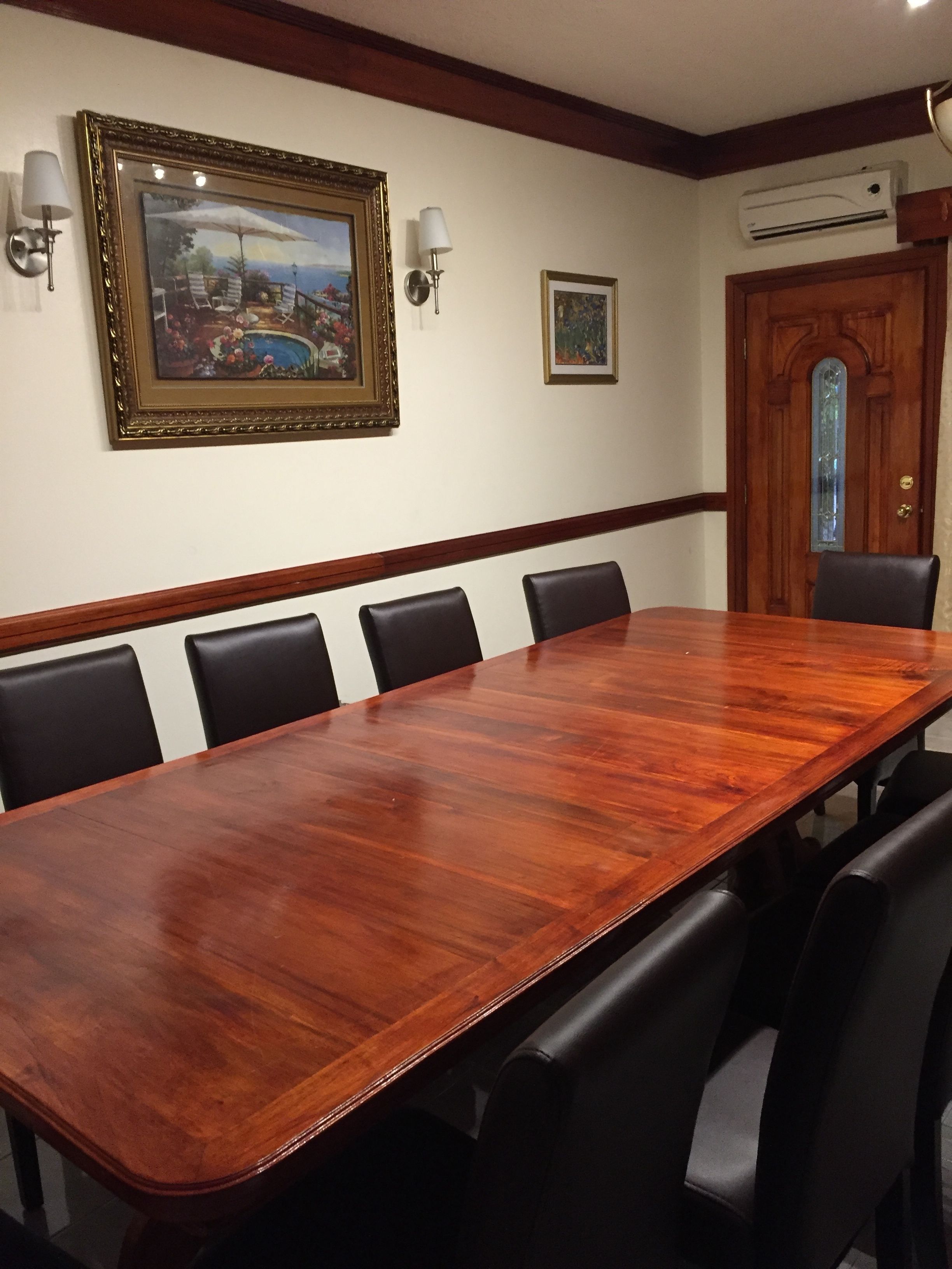 Un salón de conferencias con una larga mesa de madera, sillas negras, obras de arte enmarcadas, lámparas fijadas a la pared y un equipo de aire acondicionado.