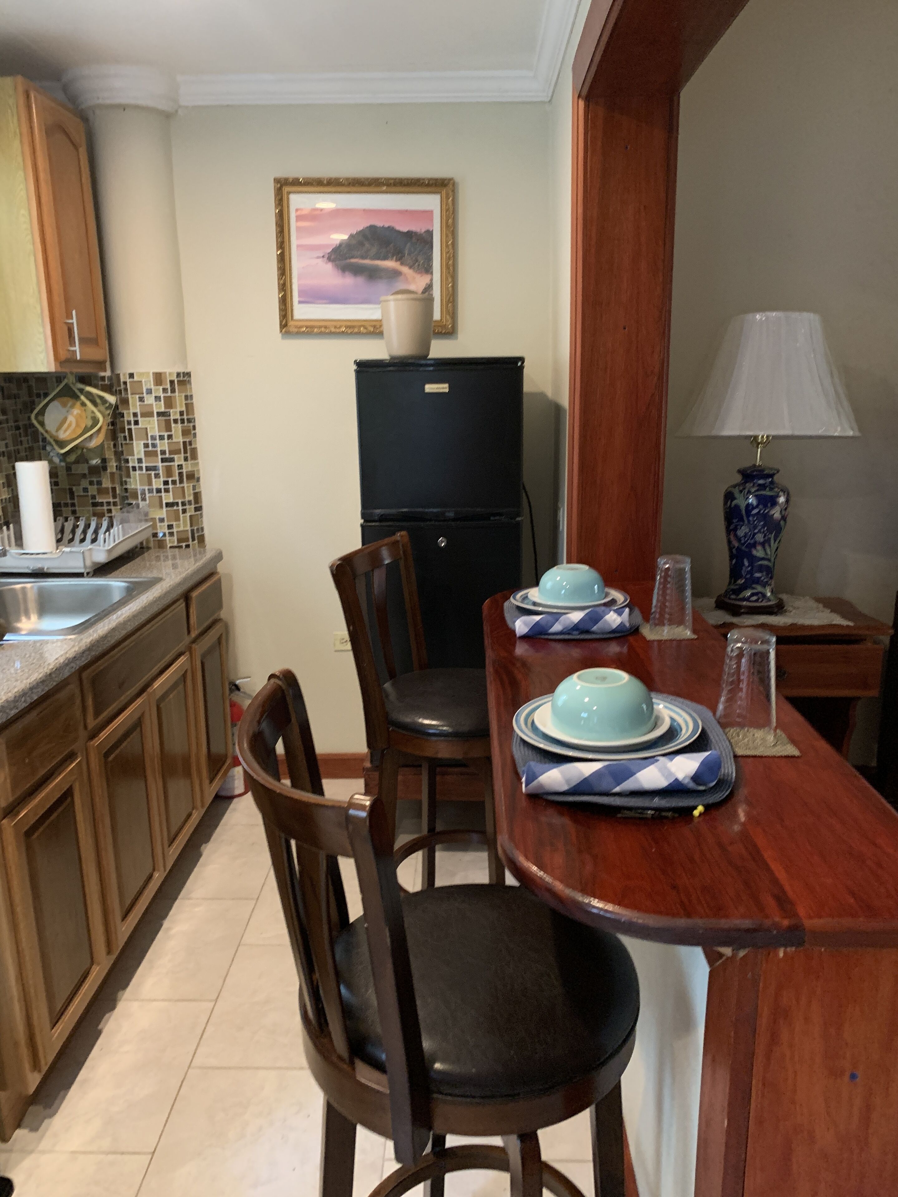 Un coin repas avec une table en bois dressée pour deux personnes, un réfrigérateur noir et une lampe décorative.