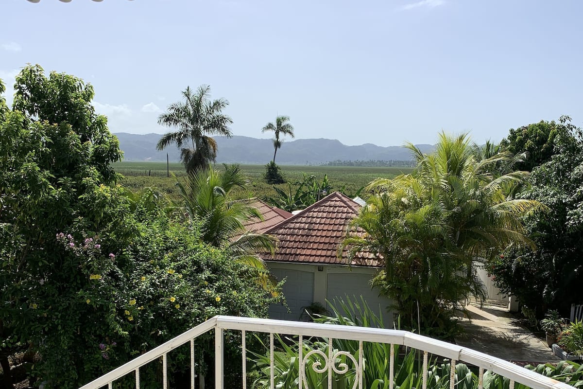 Un balcón con vistas a una casa, abundante vegetación y montañas lejanas.