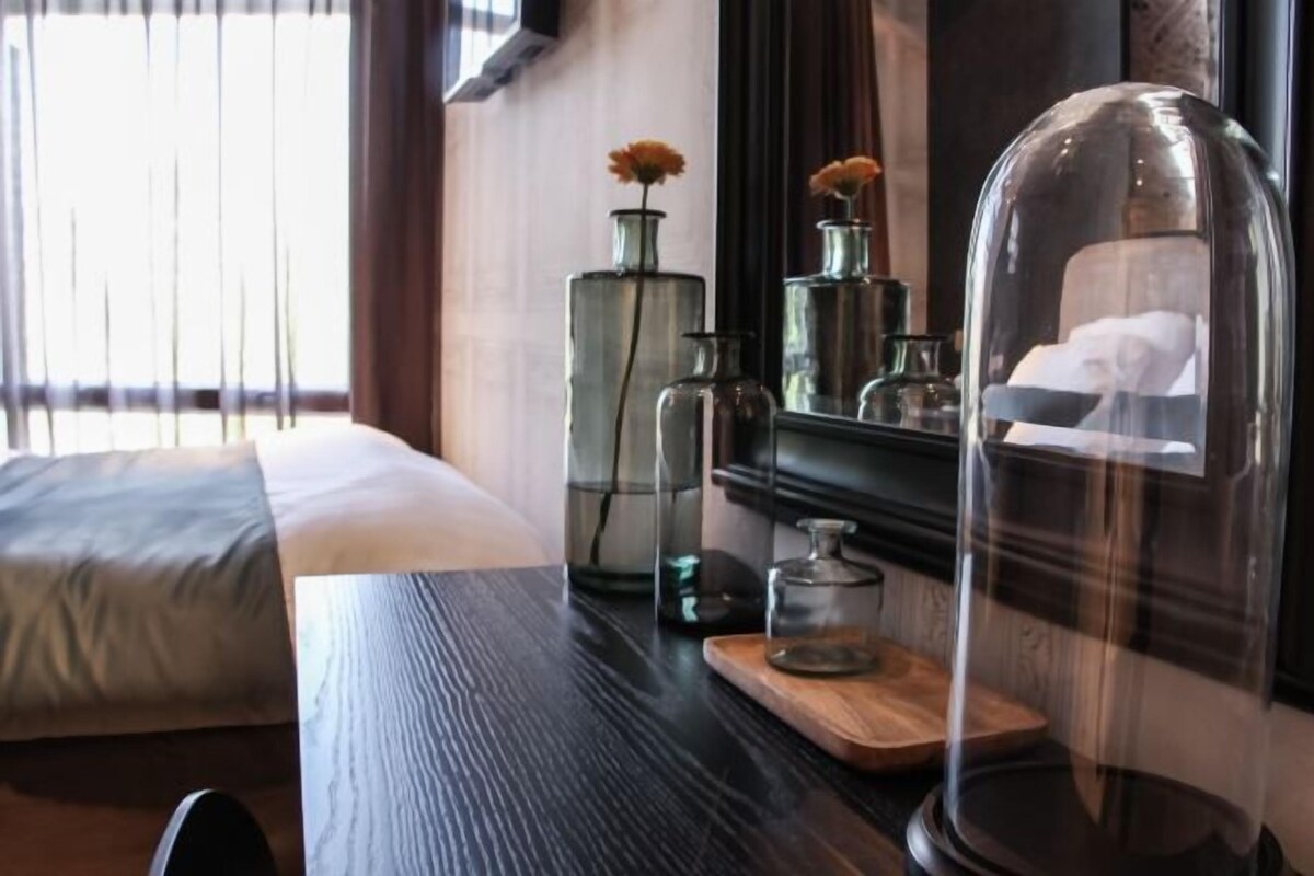 A hotel room with a bed, a wooden table with decorative items, and a window with blinds.