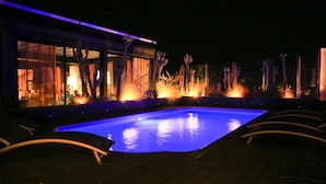 A nighttime swimming pool area with a clear pool, surrounding plants, and a building with large windows. - Medina Loft & Spa (Marrakech)