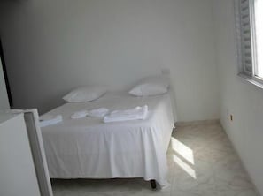 A small, clean room with a single bed, white bedding, and a window providing natural light.