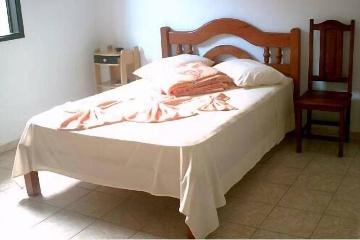 Ein Einzelbett mit Holz-Kopfteil, ein Nachttisch und ein Stuhl in einem Zimmer mit Fliesenboden.