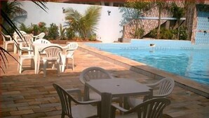 Uma área à beira da piscina com mesa e cadeiras, cercada por um pátio com piso de pedra.