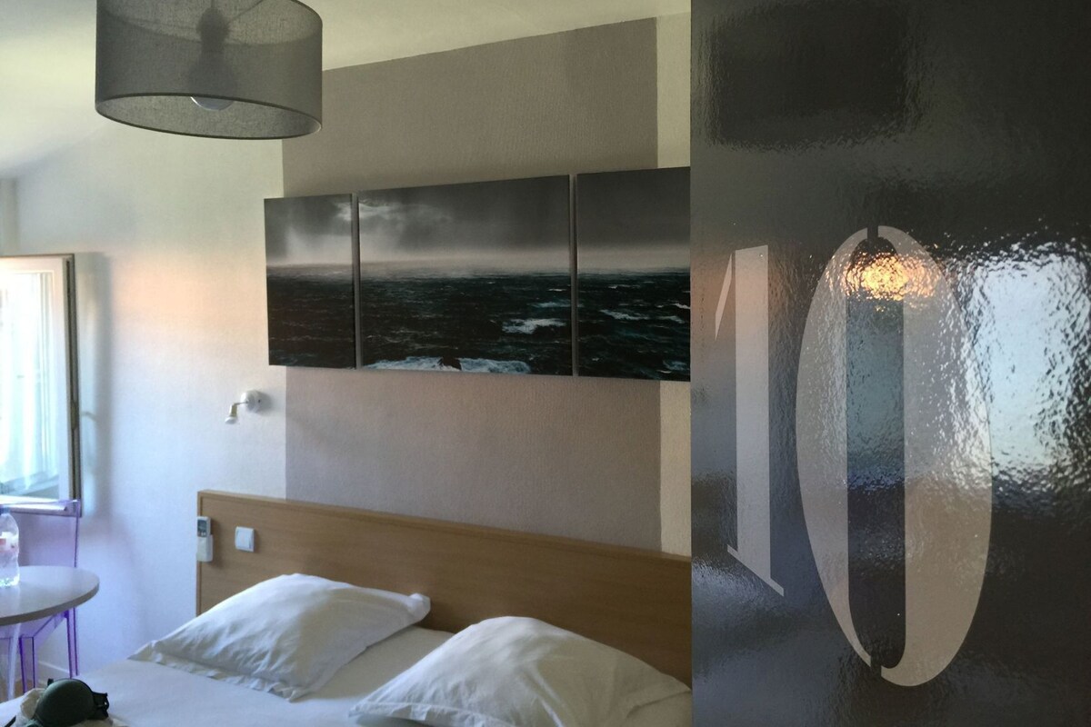 A hotel room with a bed, a round table, a wall-mounted artwork, and a hanging light fixture.