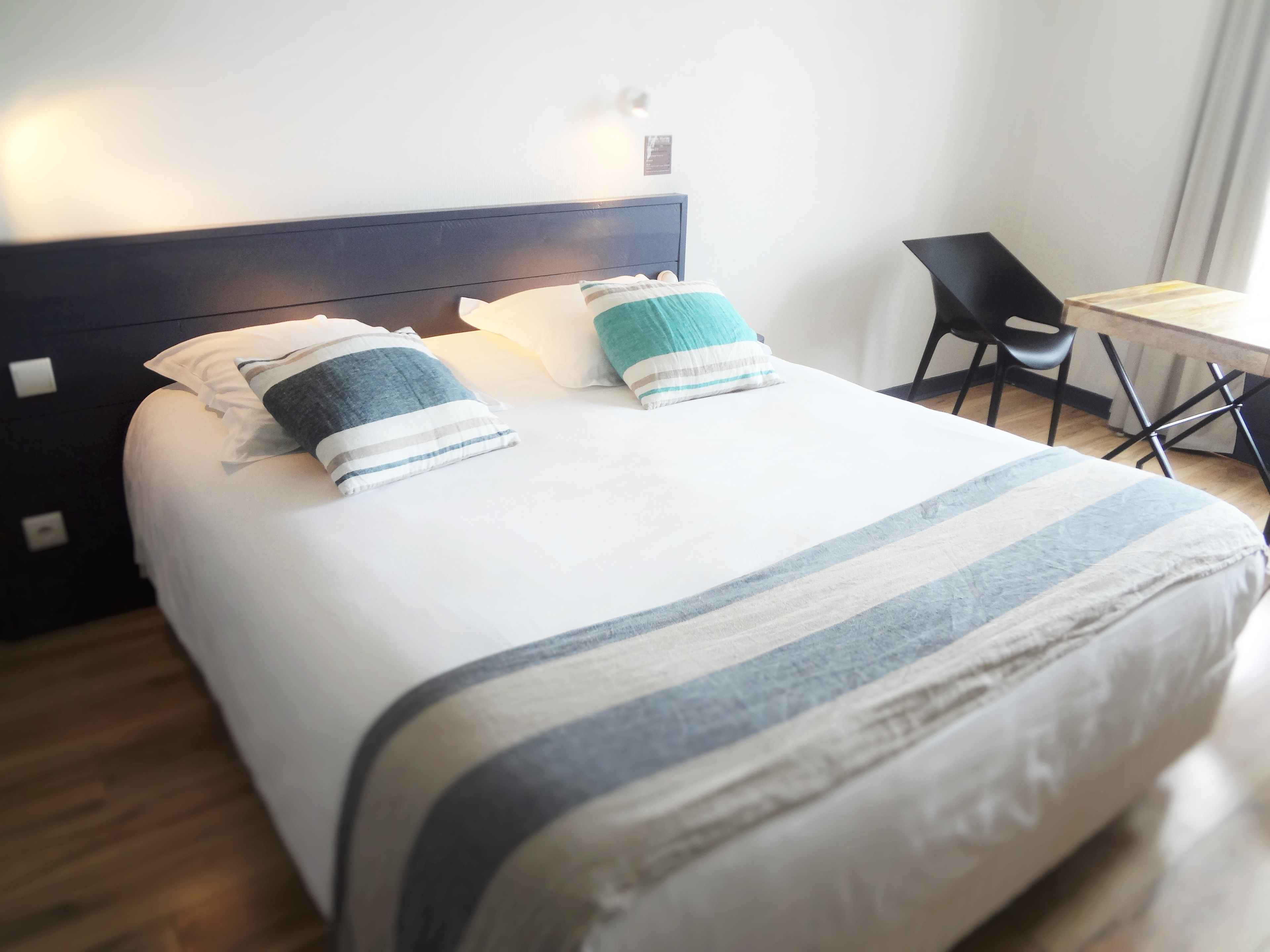 Cama bem arrumada com roupa de cama listrada, cabeceira escura e uma pequena mesa com uma cadeira ao lado.