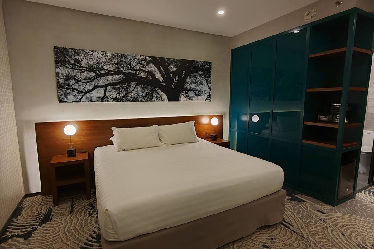 A hotel room with a large bed, a nightstand with a lamp, a wall-mounted artwork of a tree, and a teal wardrobe.