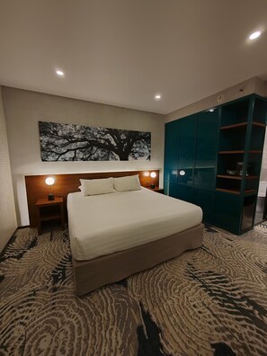 A hotel room with a large bed, a nightstand with a lamp, a wall-mounted artwork of a tree, and a teal wardrobe. - Isla Verde Hotel (San José)