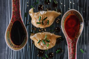Una cuchara decorativa con diseño estampado, un par de dumplings adornados con cebolletas y semillas de sésamo, y un cuenco de salsa roja.