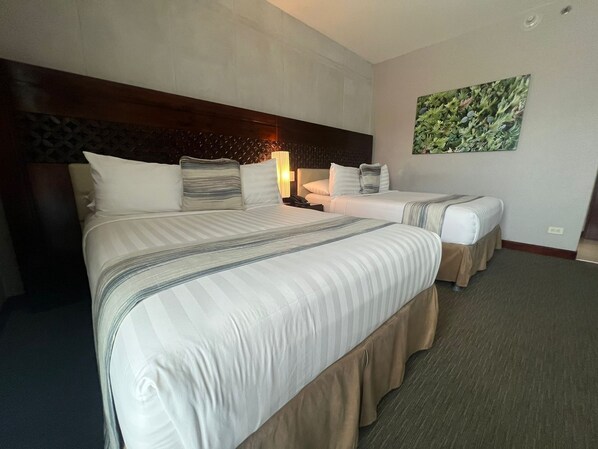 A hotel room with two beds, a headboard, a nightstand, a lamp, and a wall-mounted artwork. - Isla Verde Hotel (San José)