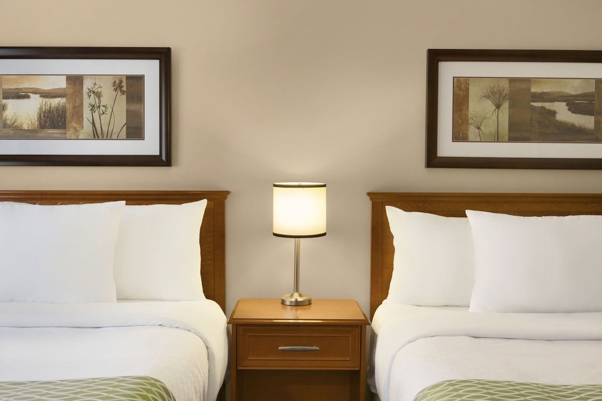 Una habitación de hotel con dos camas, una mesita de noche con lámpara y dos cuadros enmarcados en la pared.