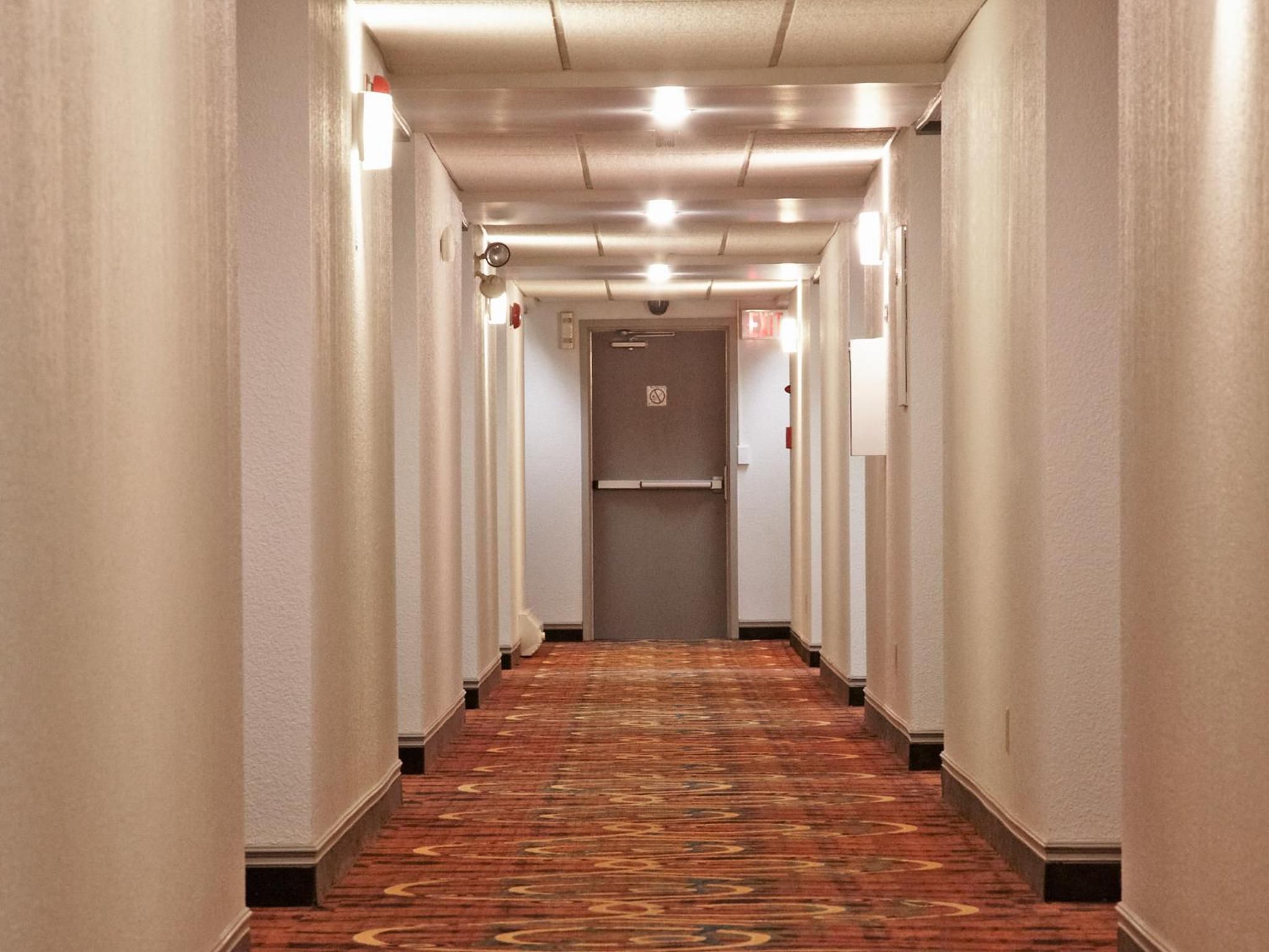Corredor de hotel com piso em carpete, paredes brancas e uma porta no final.