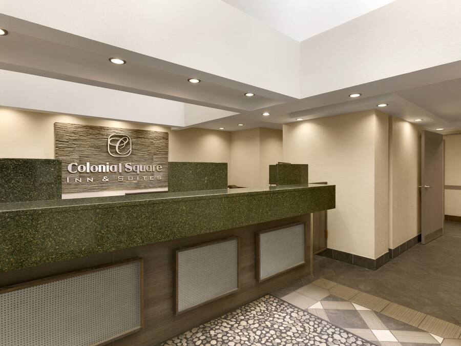 A reception desk with a sign reading "Colonial Square Inn & Suites," a modern design with a green countertop, recessed lighting, and a patterned floor.
