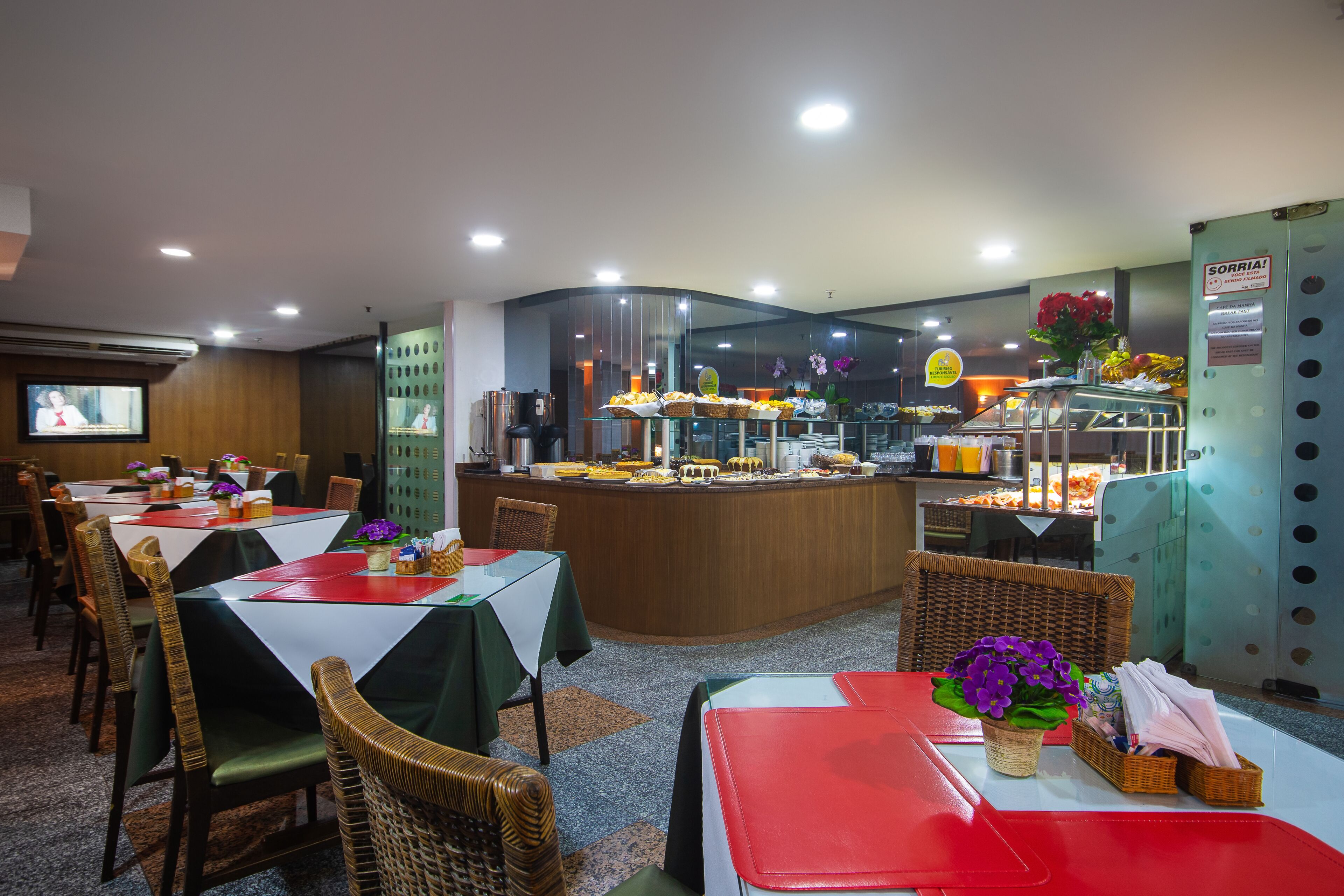 A restaurant with multiple tables and chairs, a counter with food displays, and a television screen.