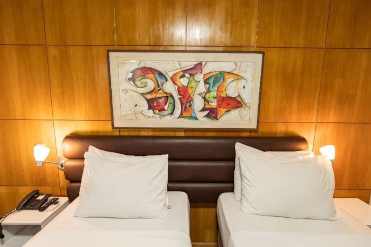 A hotel room with a wooden headboard, two beds with white linens, a framed abstract artwork above the headboard, and bedside lamps.
