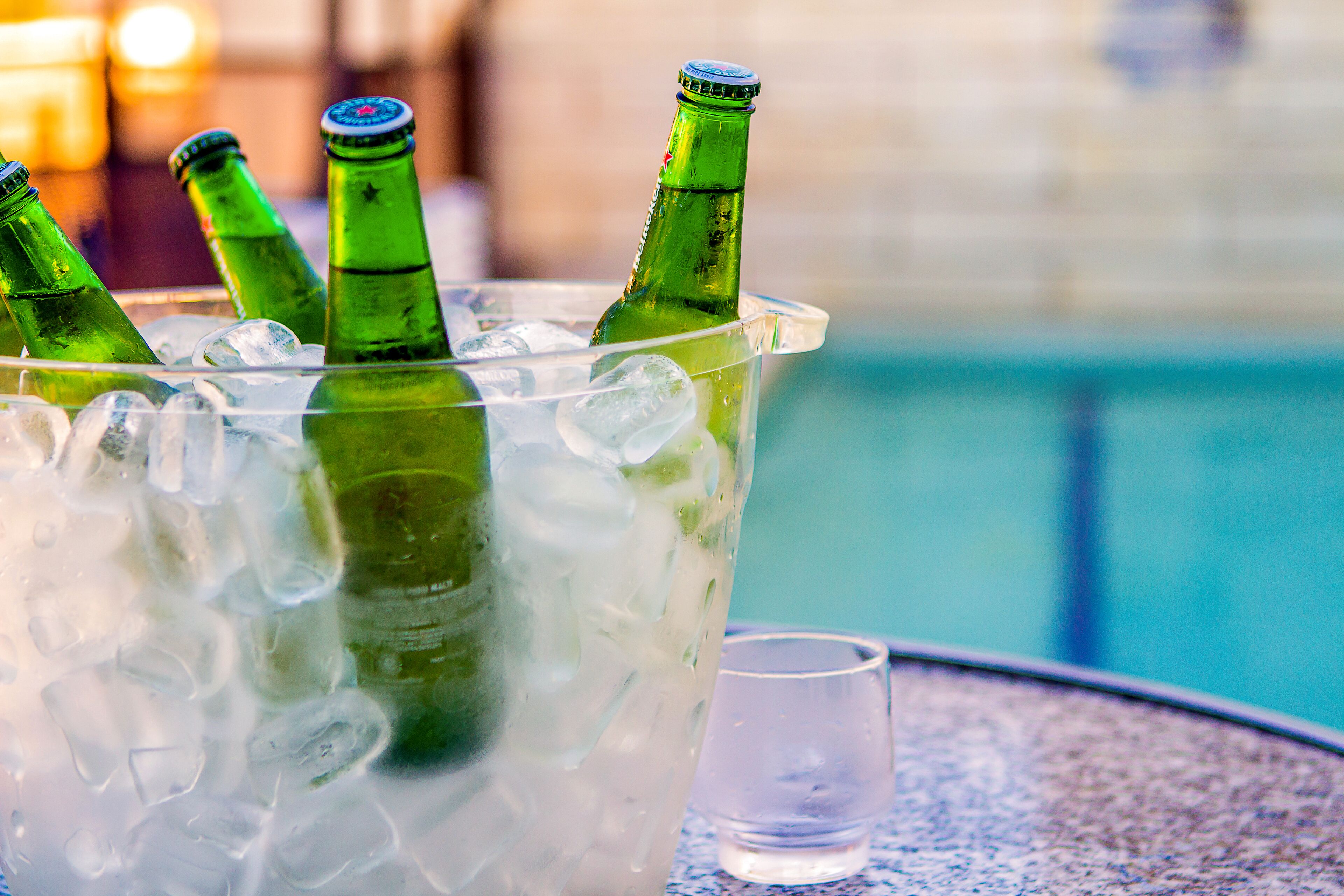 A clear glass container with ice and three green bottles, placed on a table near a pool.