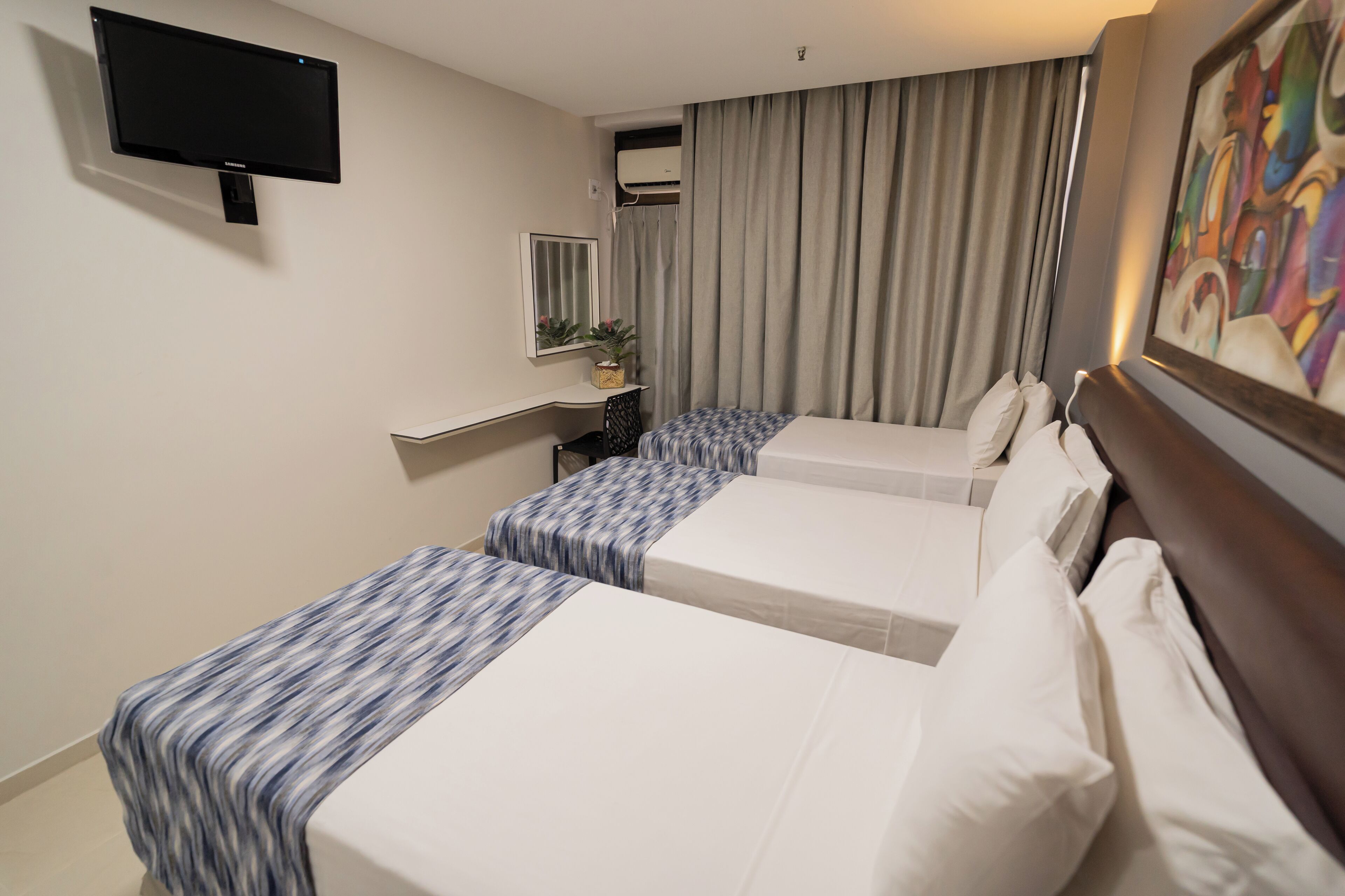 A hotel room with two beds, a flat-screen TV mounted on the wall, a desk with a chair, a window with curtains, and a painting on the wall.