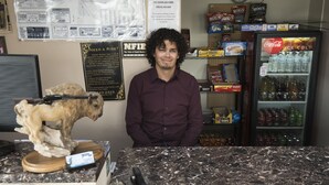 A man standing behind a counter with a dog figurine, a Coca-Cola refrigerator, and various snacks. - Travel Inn (Laramie)