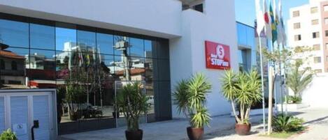 A modern building with a glass facade, a red sign with white text, and several potted plants in the foreground.