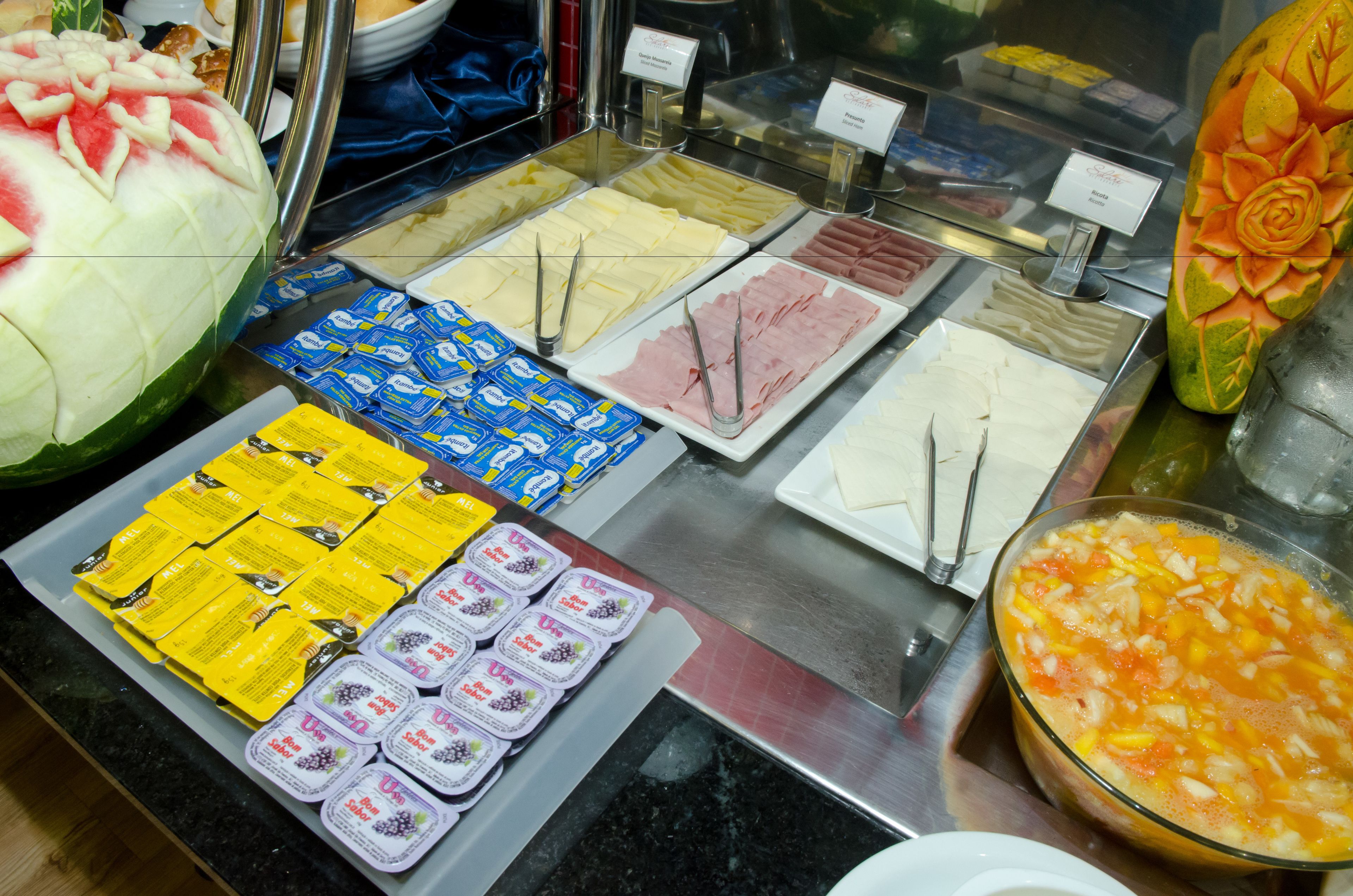A buffet with various food items including a watermelon, cheese, ham, and a bowl of fruit salad.