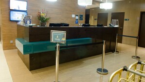 A hotel reception area with a glass counter, two monitors, a potted plant, and a sign indicating room rates. - Ímpar Suítes Expominas (Belo Horizonte)