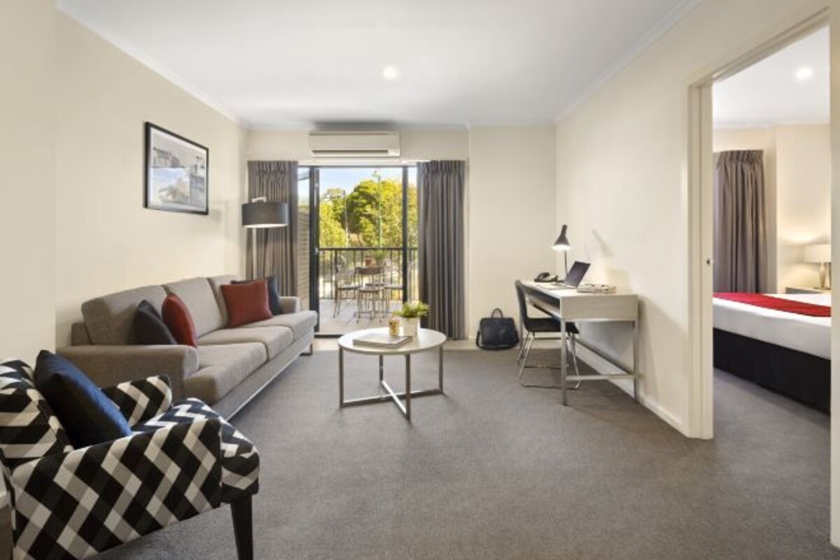A modern hotel room with a sofa, a desk, a small round table, and a balcony with a view of greenery.