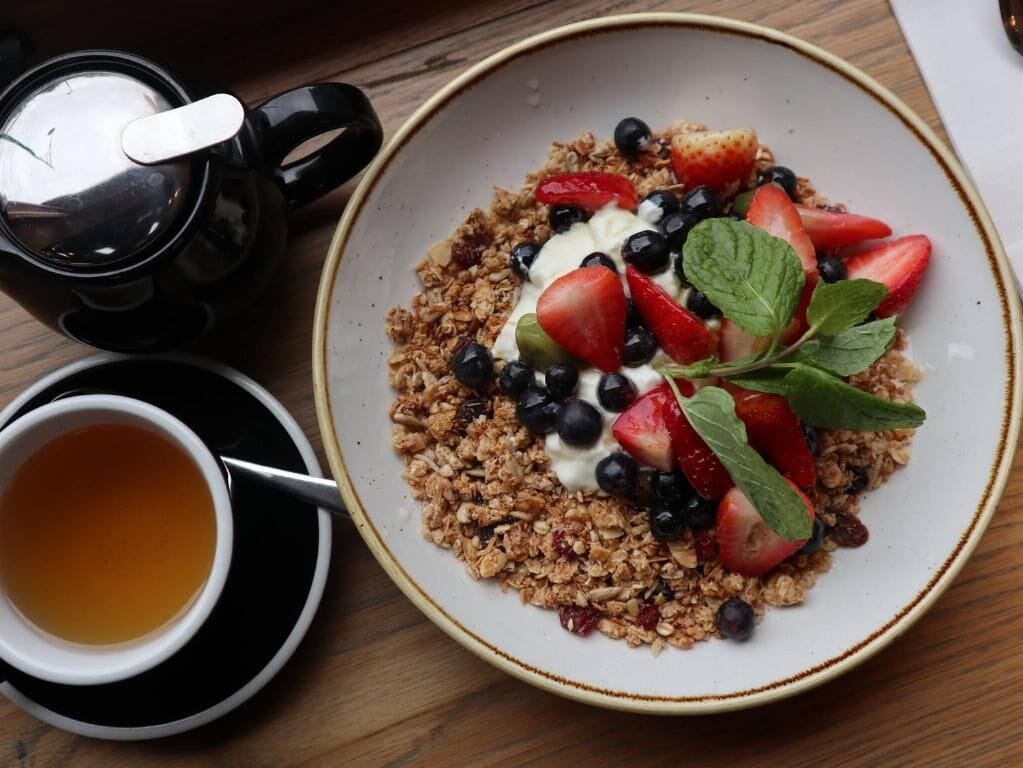Una mesa de desayuno con un plato de granola cubierto de yogur, fresas, arándanos y hojas de menta, acompañado de una taza de té y una cafetera.