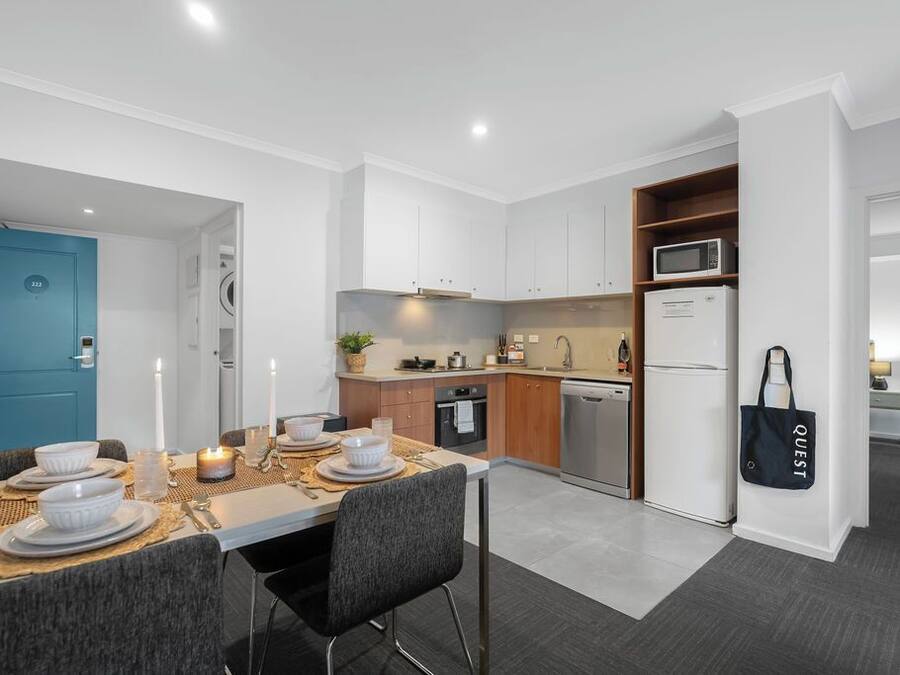 A modern kitchen with white cabinets, a microwave, and a refrigerator. There is a dining area with a table set for two, and a blue door in the background.