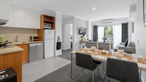 Une cuisine moderne avec des meubles blancs, un four à micro-ondes et un lave-vaisselle. On y trouve un coin repas avec une table dressée pour deux personnes, un téléviseur fixé au mur et un canapé à l’arrière-plan.