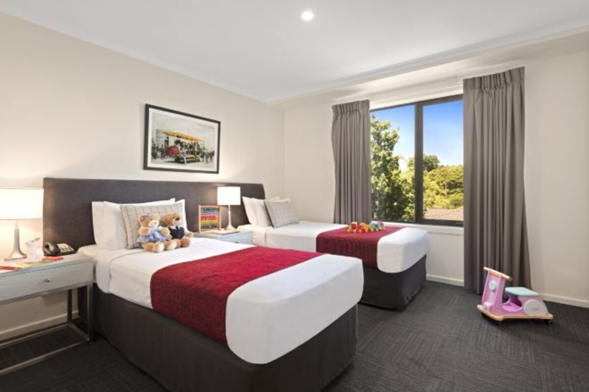 A hotel room with two beds, a desk, a chair, a window with curtains, and a framed picture on the wall.