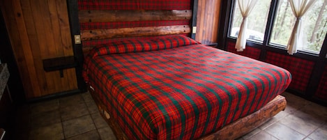 A wooden bed with a plaid bedspread in a room with wooden walls and a window with curtains.