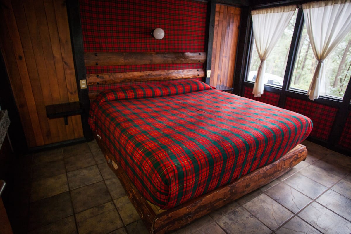 Een houten bed met een geruite sprei in een kamer met houten wanden en een raam met gordijnen.