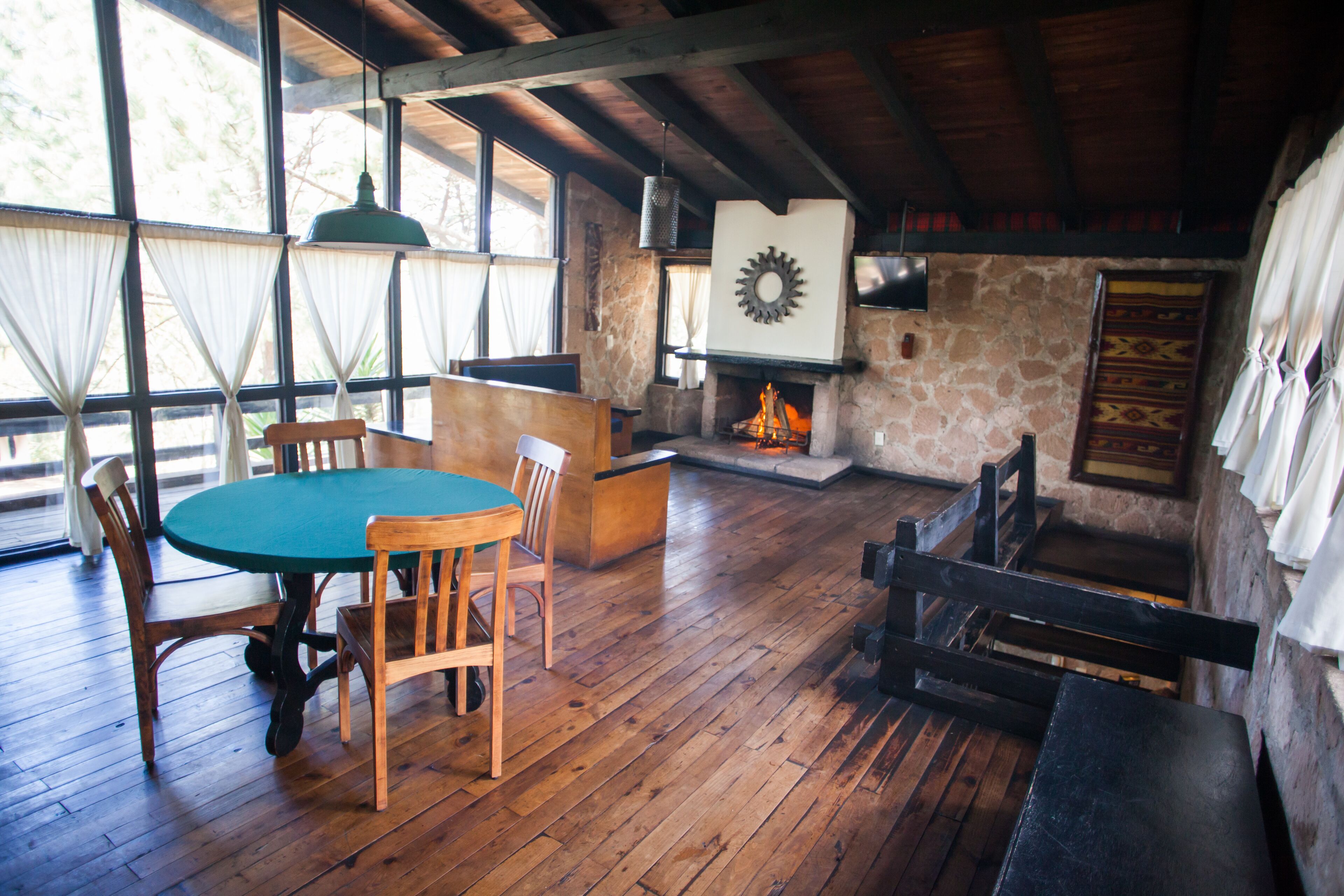 A rustic interior with a wooden dining table, chairs, a stone fireplace, and a large window with curtains.