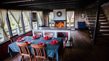 A dining area with a table set for four, wooden chairs, a fireplace, and a view through large windows.