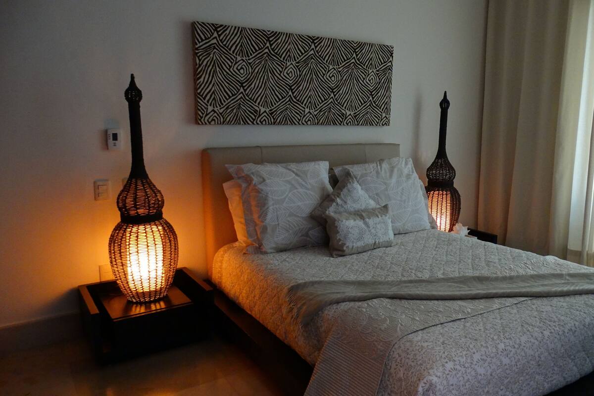 Una cama bien tendida con un cabecero estampado, dos lámparas de noche y una obra de arte colgada en la pared.