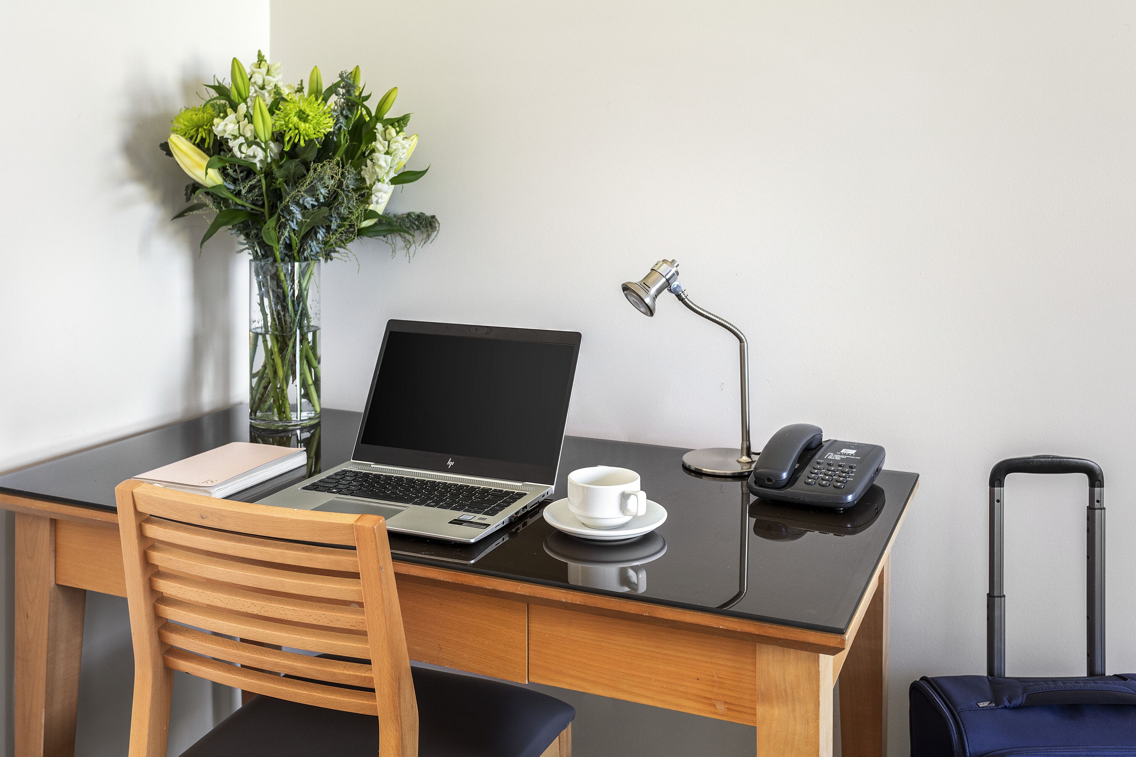 A hotel workspace with a laptop, a cup, a phone, a vase with flowers, and a suitcase.