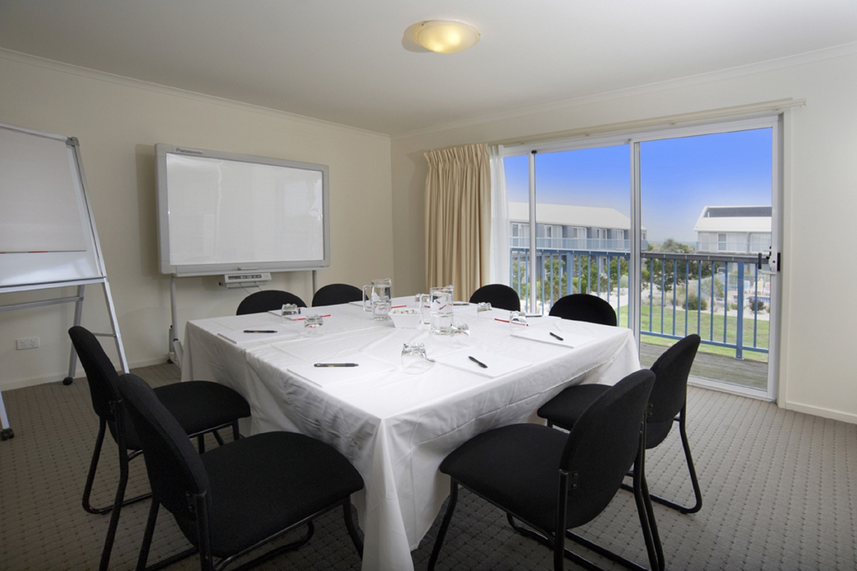 A conference room with a white table, black chairs, a whiteboard, and a view of a building through large windows.