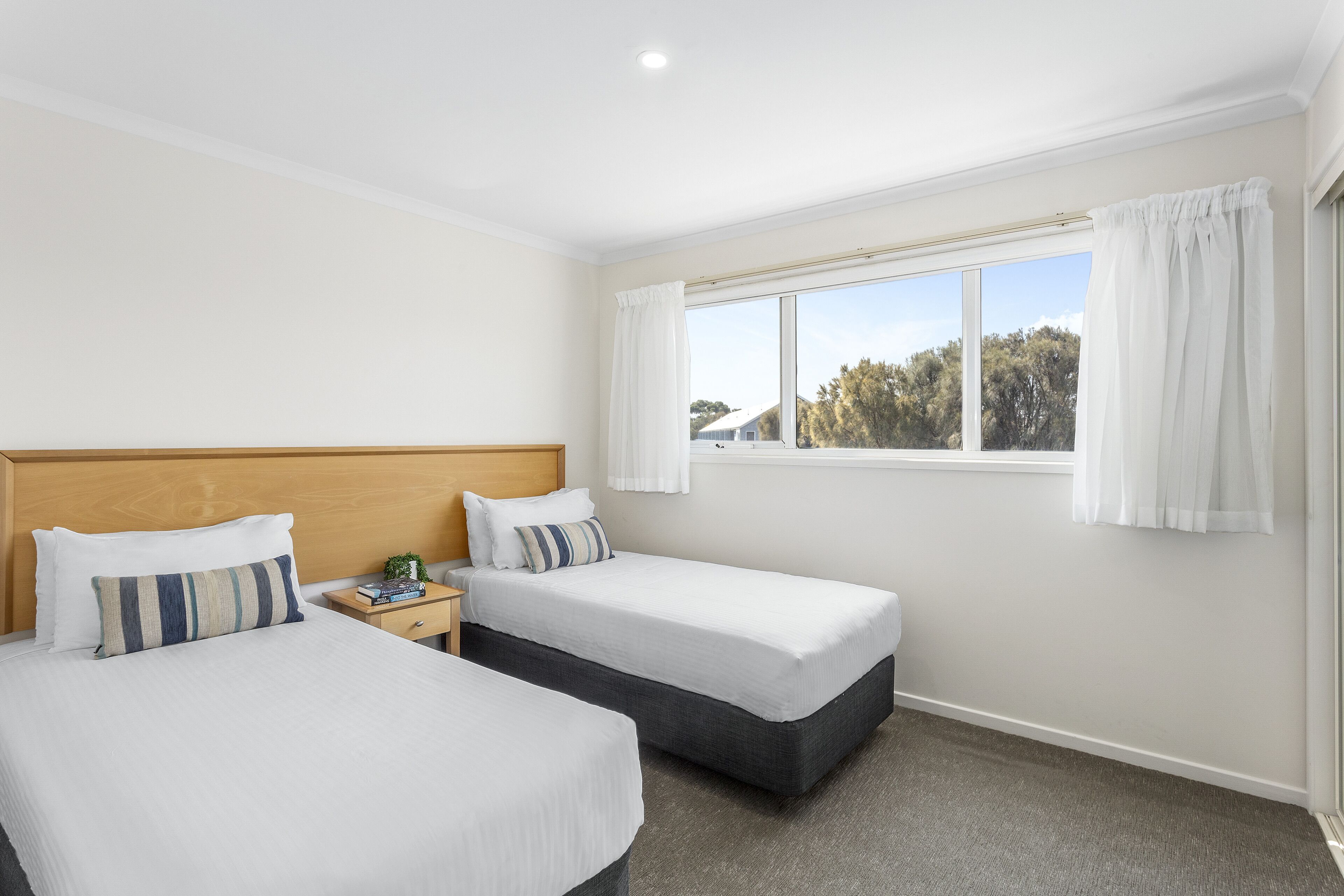 A hotel room with two single beds, a wooden headboard, a nightstand, and a window with curtains.