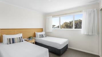 Una habitación de hotel con dos camas individuales, cabecera de madera, mesita de noche y ventana con cortinas.