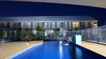 A swimming pool with a blue interior, surrounded by a fence, and a building with multiple balconies in the background.