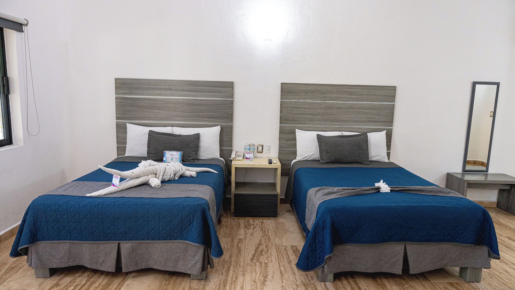 A hotel room with two beds, each with a blue quilted bedspread and grey pillows. There is a wooden nightstand between the beds with a phone and a small table with a remote control.