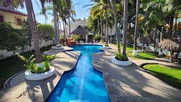 A swimming pool with a blue deck, surrounded by palm trees and a thatched-roof structure.