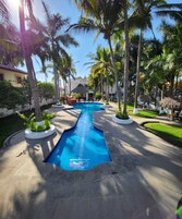 A swimming pool with a blue deck, surrounded by palm trees and a thatched-roof structure.