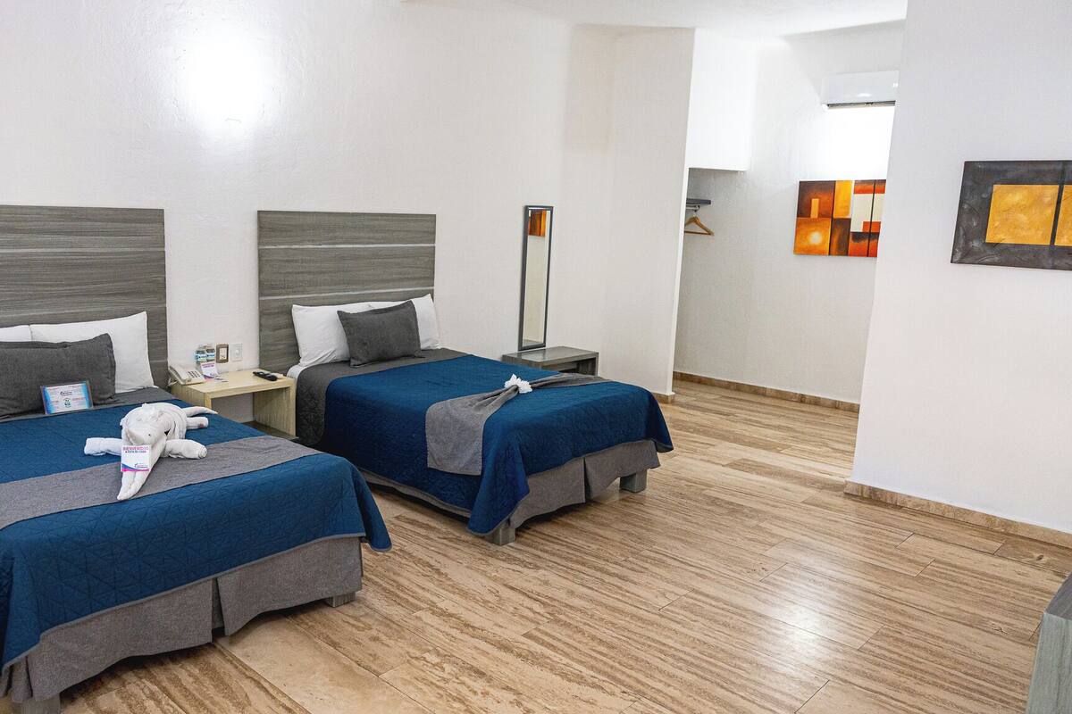 Ein Hotelzimmer mit zwei Betten, jeweils mit blauer Bettdecke und grauen Kopfteilen. An einer Seite befindet sich ein Nachttisch mit Lampe und Telefon. Der Raum hat Holzboden und einen Spiegel an der Wand.
