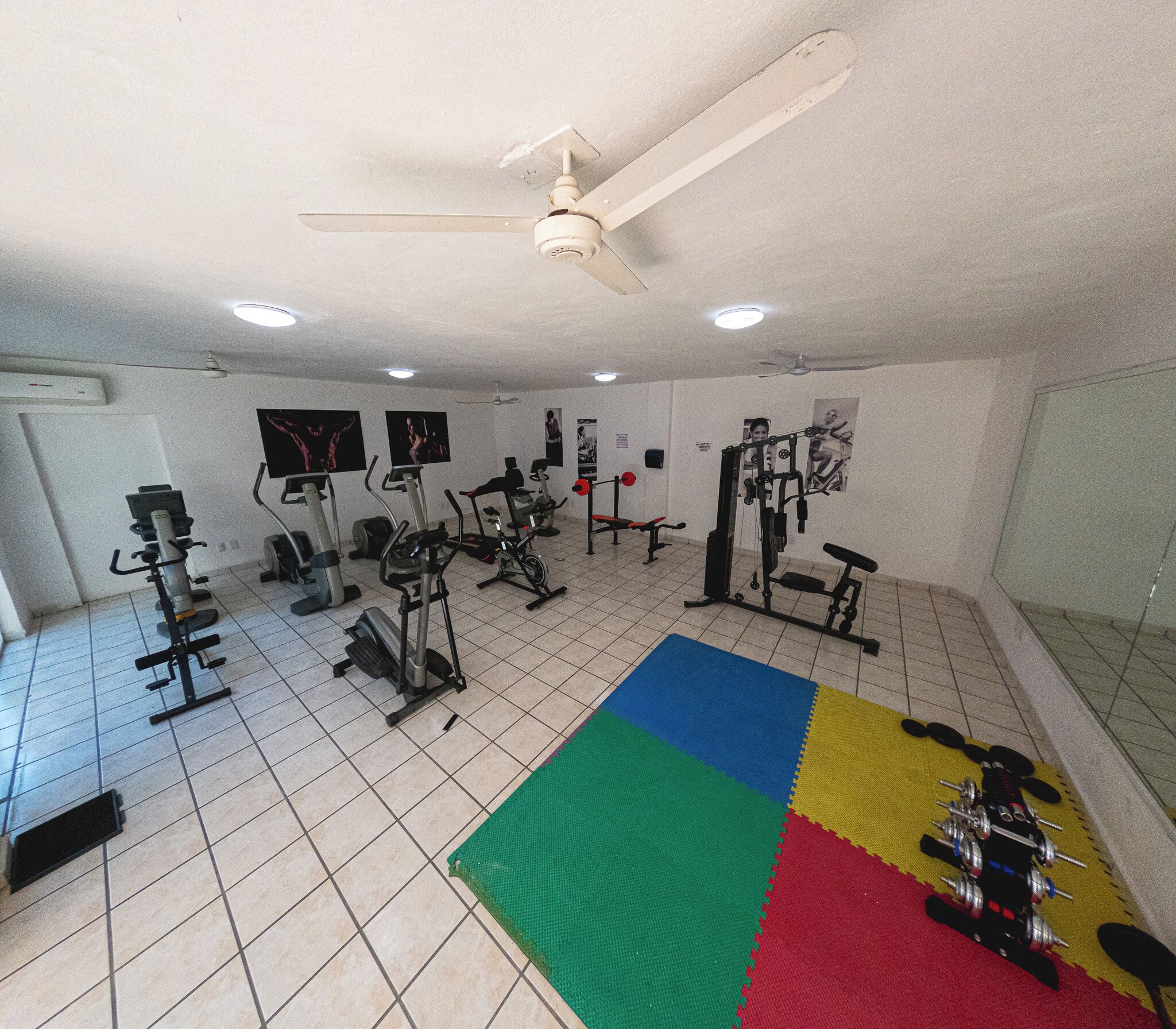 Une salle de sport équipée de divers appareils d’exercice, avec un ventilateur au plafond et un tapis coloré sur le sol.